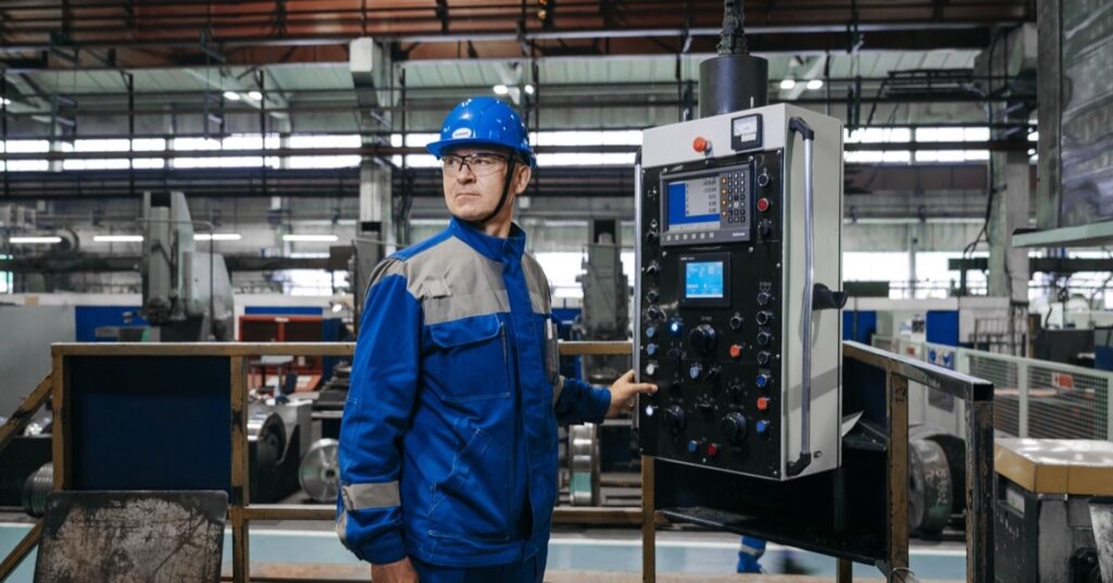 Technician in a blue safety helmet standing next to the control panel of a machine in a factory hall