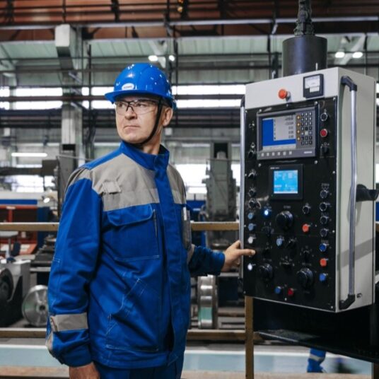 edge-case-testing Technician in a blue safety helmet standing next to the control panel of a machine in a factory hall