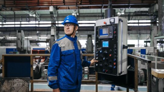 Technician in a blue safety helmet standing next to the control panel of a machine in a factory hall