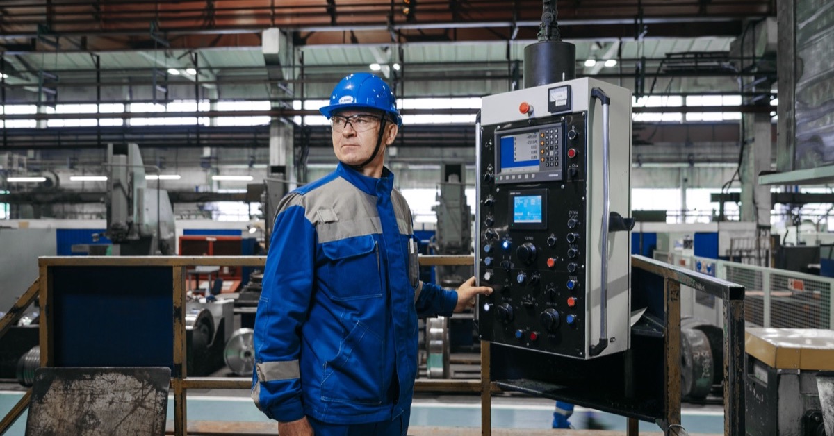 Technicus met blauwe veiligheidshelm naast het bedieningspaneel van een machine in een fabriekshal