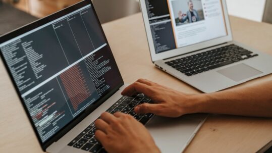 Handen typen op een laptop met terminalvensters vol rode foutregels, met een tweede laptop op de achtergrond