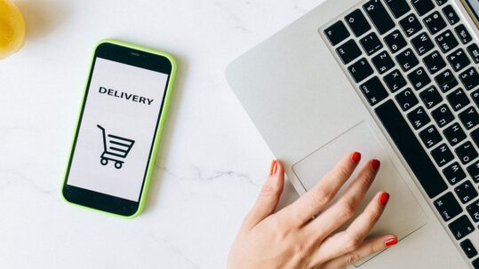 Telefoon met DELIVERY en een winkelwagen-icoon naast een hand op een laptop, op een marmeren tafel