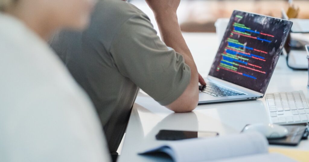 Iemand werkt aan een wit bureau op een laptop vol kleurige code, met een onscherpe collega op de voorgrond