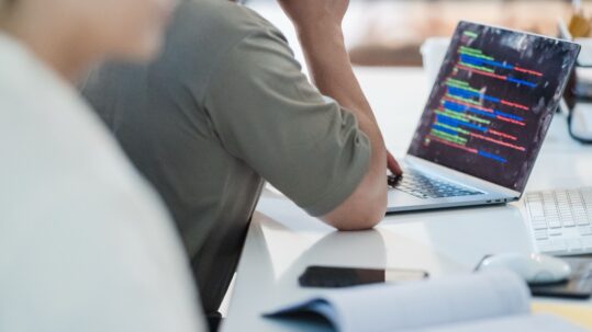 Person working at a white desk on a laptop full of colourful code, with a colleague out of focus in the foreground