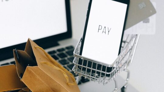 Telefoon met PAY in een mini-winkelwagentje naast een laptop, een bankpas en papieren tasjes