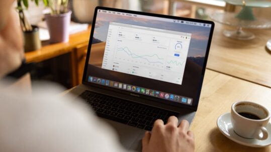 Iemand werkt op een laptop met een verkoopdashboard vol grafieken, met een kop koffie op tafel