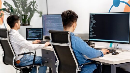 Two testers seen from behind at desks with large monitors showing code and a terminal