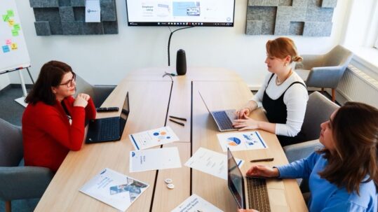 Three colleagues at a table work through laptops and documents with charts